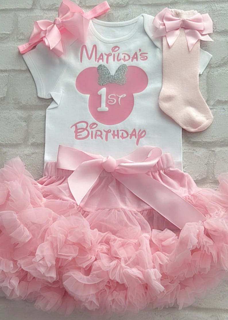 May include: A white short-sleeved bodysuit with the text Matilda's 1st Birthday and a pink mouse silhouette with a silver glitter bow. Includes a pink ruffled tulle skirt with a large satin bow, a pink headband with a bow, and pink socks with bows.