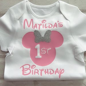 May include: A white short-sleeved bodysuit featuring a pink silhouette of a mouse head with a silver glitter bow. The text reads Matilda's 1st Birthday in pink lettering, with the number 1 in white, centered on the front of the garment.