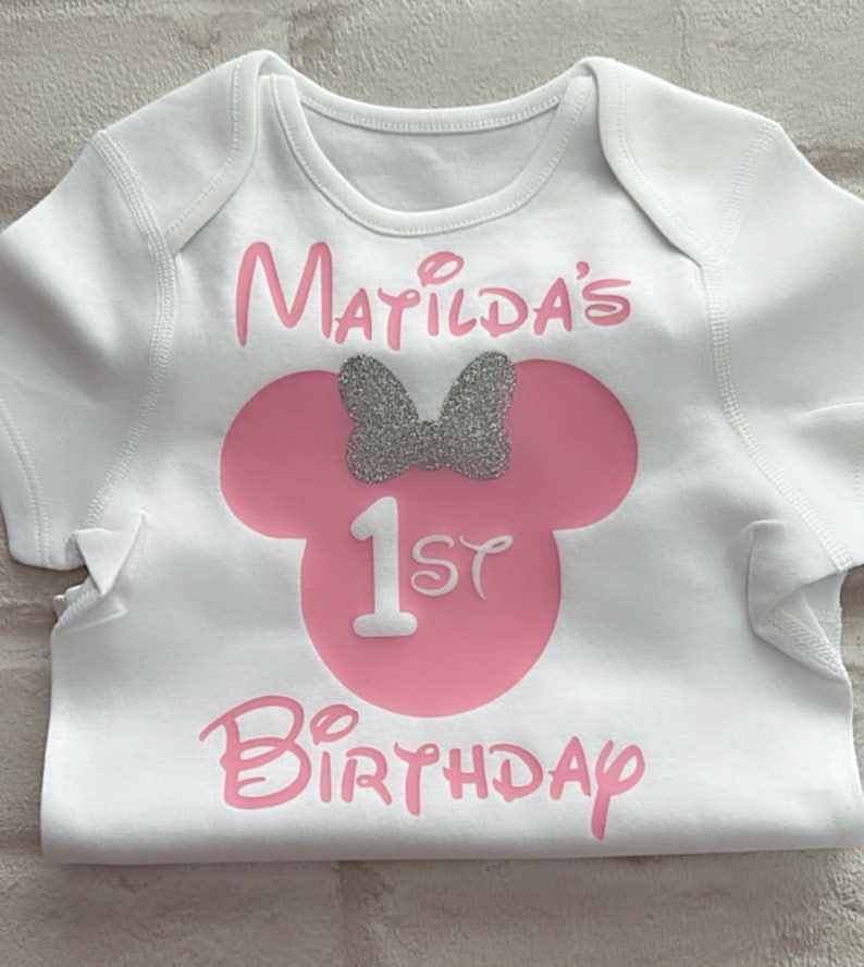 May include: A white short-sleeved bodysuit featuring a pink silhouette of a mouse head with a silver glitter bow. The text reads Matilda's 1st Birthday in pink lettering, with the number 1 in white, centered on the front of the garment.