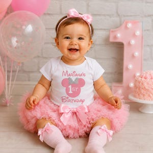 May include: A white short-sleeved bodysuit with a pink graphic featuring a mouse silhouette, a silver glitter bow, and the text Mariana's 1st Birthday, paired with a pink layered tulle tutu skirt with a bow and matching pink knee-high socks.