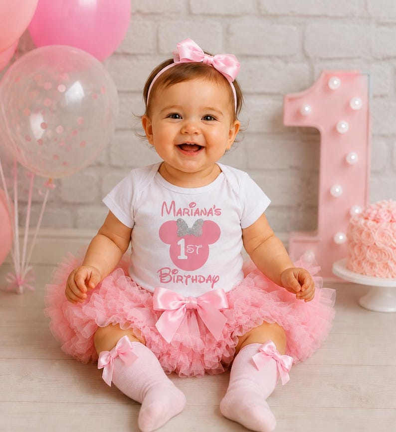 May include: A white short-sleeved bodysuit with a pink graphic featuring a mouse silhouette, a silver glitter bow, and the text Mariana's 1st Birthday, paired with a pink layered tulle tutu skirt with a bow and matching pink knee-high socks.