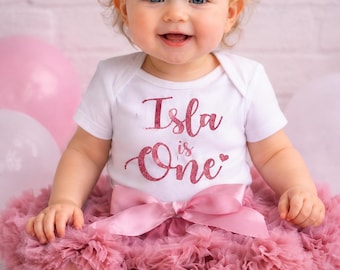 May include: A baby wearing a white onesie with the text "Isla is One" in pink glitter, paired with a pink tutu skirt, bow headband, and socks. The outfit is designed for a first birthday celebration.