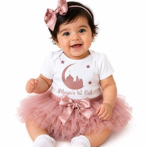 May include: A baby wearing a white onesie with a rose gold mosque and crescent moon design, the text "Alaya's 1st Eid", and a pink tutu skirt. The baby is also wearing a matching pink bow headband and a large bow at the waist.