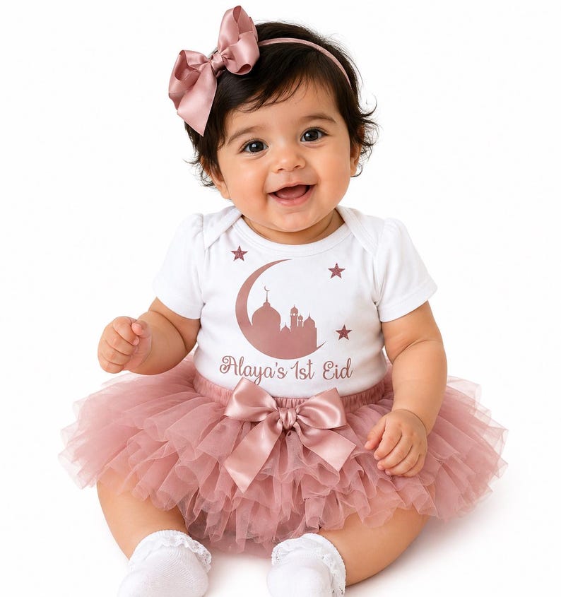 May include: A baby wearing a white onesie with a rose gold mosque and crescent moon design, the text "Alaya's 1st Eid", and a pink tutu skirt. The baby is also wearing a matching pink bow headband and a large bow at the waist.