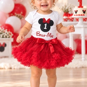 May include: A white short-sleeved bodysuit featuring a black glittery mouse ear silhouette with a red bow and the number 1, with the name Bella-Mae printed in red below. It is paired with a voluminous red tulle tutu skirt with a satin bow at the waist.