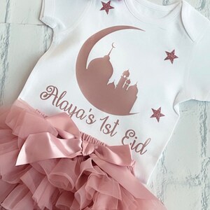 May include: A white baby outfit with a pink bow headband. The outfit includes a onesie with a mosque and crescent moon design, the text "Alaya's 1st Eid", and a ruffled diaper cover. Pink glitter stars are also on the onesie.
