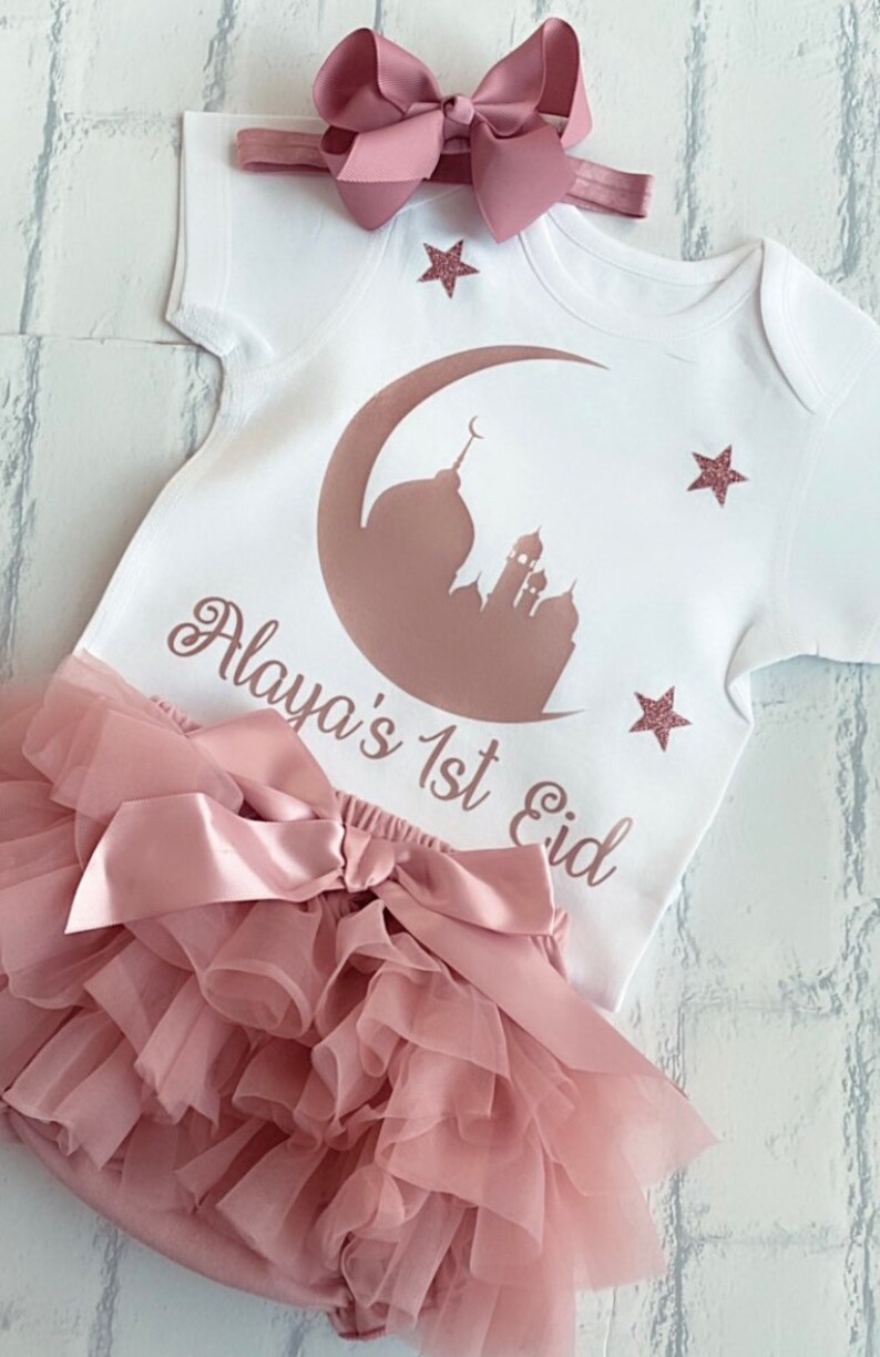 May include: A white baby outfit with a pink bow headband. The outfit includes a onesie with a mosque and crescent moon design, the text "Alaya's 1st Eid", and a ruffled diaper cover. Pink glitter stars are also on the onesie.
