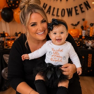 May include: A white long-sleeve bodysuit with the orange text "The cutest little pumpkin in the patch" and two small pumpkin illustrations, paired with a black tulle skirt, a black headband with a bow, and black ruffled socks.