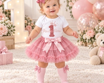 May include: A white short-sleeved bodysuit with a glittery pink crown and the number 1 above the name Amelia Rose, paired with a dusty pink layered tulle tutu skirt featuring a satin bow, a matching headband with a large bow, and knee-high socks.