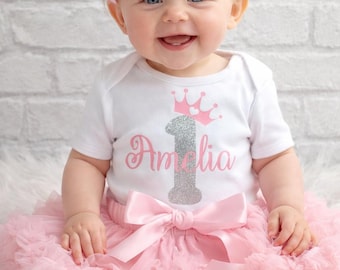 May include: A baby wearing a pink tutu, a white onesie with a pink and silver "1" and crown design, and a pink headband with a bow. The onesie has the name "Amelia" printed on it. The baby is smiling.
