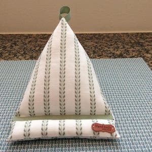 May include: A white and green striped pillow with a green ribbon loop on top. The pillow is shaped like a pyramid and has a brown label that says "Handmade with Love".