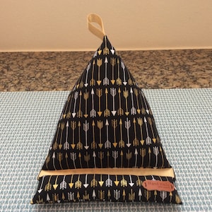 May include: A black triangular pillow with a gold and white arrow pattern. The pillow has a tan loop handle and a tan fabric bottom. The pillow has a brown leather tag that says "Handmade with love".