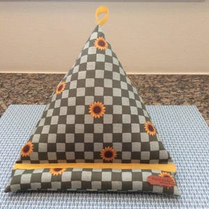 May include: A triangular fabric tablet stand featuring a gray and dark gray checkered pattern with yellow sunflower prints. The stand includes a yellow fabric strip along the base, a yellow hanging loop at the top, and a small brown tag labeled Hand made.