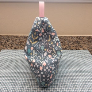 May include: A fabric bag with a floral pattern in shades of blue, pink, and yellow. The bag has a pink ribbon handle. The bag is sitting on a blue and white patterned mat.