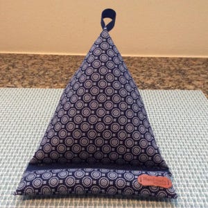 May include: A blue and white patterned triangular pillow with a blue ribbon loop at the top. The pillow is on a light gray textured surface.