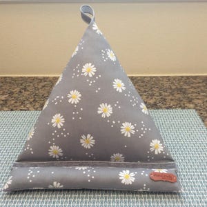 May include: Gray triangular book stand with a daisy pattern. The fabric features white daisies with yellow centers and small white dots. A small brown tag with text is attached to the bottom. A loop is at the top for easy carrying.