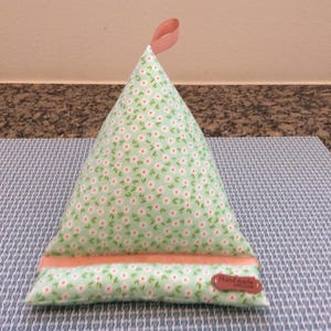 May include: A green and white floral patterned pillow with a peach colored top and a brown label that says "Handmade" on it. The pillow is shaped like a triangle and is sitting on a blue and white patterned surface.