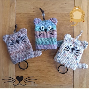 Op de afbeelding: Drie gehaakte sleutelhangers in de vorm van katten met verschillende garenkleuren. De sleutelhangers zijn bruin, blauw en wit met zwarte ogen en snorharen. Elke sleutelhanger heeft een metalen ring.