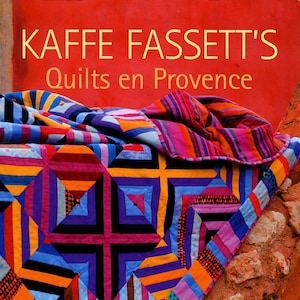 May include: A book cover featuring a colorful quilt made of various fabrics in shades of orange, red, purple, blue, and yellow. The quilt is draped over a textured red wall. The title of the book is "Kaffee Fassett's Quilts en Provence" and the subtitle is "20 designs from Rowan for patchwork and quilting".
