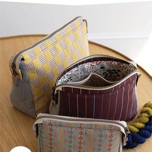 Può includere: Tre pochette in tessuto di diverse dimensioni con chiusure a zip. La più grande ha un motivo a scacchi giallo e bianco. La pochette centrale è bordeaux con strisce verticali. La più piccola è grigia con un motivo geometrico. Testo: "PLOVER POUCH" e "3 misure da cucire!"