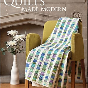Op de afbeelding: Een boek getiteld Quilts Made Modern door Weeks Ringle en Bill Kerr met een quilt met verticale strepen in tinten blauw, groen, paars en wit gedrapeerd over een gele fauteuil, met een witte vaas met witte bloemen in de buurt.