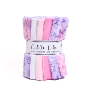 May include: A roll of five 20-inch precut fabric strips in shades of purple, pink, and white. The fabric is a soft, plush cuddle fabric. The label reads "Cuddle Cake 5 Precut 20" Strips Luxe Cuddle & C3"