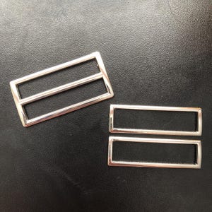 Pode incluir: Duas fivelas retangulares prateadas com uma barra central. As fivelas, feitas de metal brilhante, estão sobre um fundo escuro. Estas fivelas são provavelmente para cintos, bolsas ou outros acessórios, e medem cerca de 5 cm de comprimento.