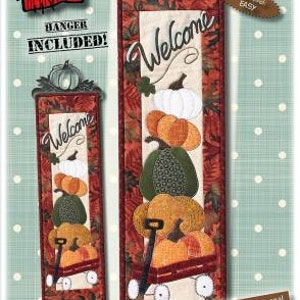 Könnte beinhalten: Willkommen Wagen Quiltmuster mit einem roten Wagen, einem Stapel Kürbisse und einem Willkommensschild. Das Muster ist 15,2 cm x 55,9 cm groß und beinhaltet einen Aufhänger. Der Schwierigkeitsgrad ist leicht.