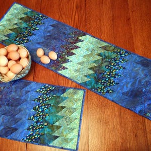 Może przedstawiać: Dwa błękitne i zielone pikowane bieżniki stołowe z wzorem diamentowym. Większy bieżnik ma szerokość 46 cm, a mniejszy 30 cm.