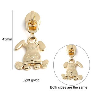 May include: Gold-colored zipper pull with a cartoon dog design. The dog is sitting on its hind legs with its front paws outstretched. The zipper pull is 43 mm long.