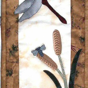 May include: A vertical fabric wall hanging featuring a quilted scene with two large grey dragonflies, brown cattails, and green lily pads on a blue water section. The border is a brown fabric with a leaf pattern and small dragonfly motifs.