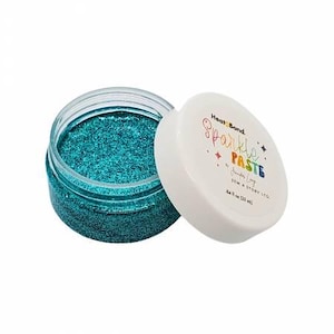 May include: An open jar of teal glitter paste with a white lid. The lid is off to the side, revealing the glitter inside. The lid has the words "Heat Bond Sparkle Paste" and "Sew a Story LTD" printed on it. The jar contains 25 ml of glitter paste.