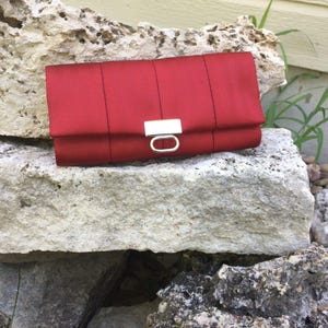 May include: A red clutch with a rectangular shape and a silver-colored clasp. The clutch is made of a smooth, possibly satin-like material. The bag is resting on a light-colored rock formation.