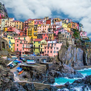 Pode incluir: Uma vila costeira colorida com edifícios em tons de amarelo, rosa e vermelho, situada numa falésia rochosa. Pequenos barcos estão atracados perto da costa, com o mar azul e o céu como pano de fundo. Uma cena pitoresca.