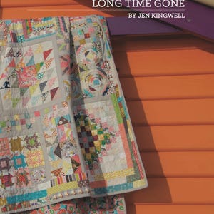 Könnte beinhalten: Ein bunter Quilt mit verschiedenen geometrischen Mustern, darunter Dreiecke und Quadrate, hängt an einer orangefarbenen Wand. Der Quilt weist eine Mischung aus pastellfarbenen und leuchtenden Stoffen auf. Der Text an der Wand lautet "LONG TIME GONE" von Jen Kingwell.