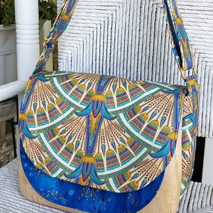 May include: A colorful shoulder bag with a flap closure. The bag features a patterned design in shades of blue, gold, and teal. The flap and strap are adorned with the same pattern. The base of the bag is a solid tan color.