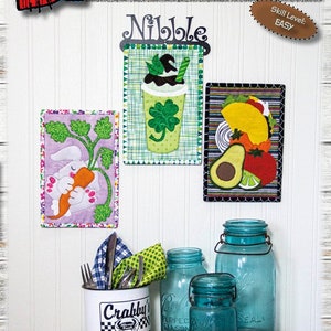 Könnte beinhalten: Eine Präsentation von Stoffkunstwerken zum Thema Frühling, darunter ein Hase mit einer Karotte, ein grünes Getränk mit einem Kleeblatt und ein Taco mit Avocado. Der Text lautet "SPRING NIBBLERS" und "EACH FINISHES: 15 cm x 23 cm". Blaue Einmachgläser und eine "Crabby"-Tasse sind ebenfalls zu sehen.