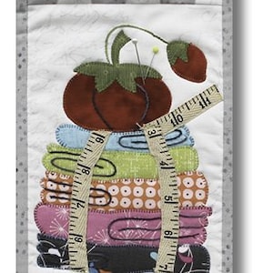 May include: A colorful quilted wall hanging featuring a stack of fabric scraps tied with a measuring tape and topped with a red and green fabric strawberry. The fabric scraps are decorated with polka dots, stripes, and other patterns. The wall hanging is framed with a black metal hanger and has a white and gray checkered background.