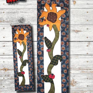 Könnte beinhalten: Ein anfängerfreundliches Quiltmuster für einen Sonnenblumen-Wandbehang. Das Muster enthält eine Sonnenblume mit gelber Mitte, braunen Blütenblättern und grünen Blättern. Die Sonnenblume befindet sich auf einem weißen Hintergrund mit einem blauen und weißen gemusterten Rand. Das Muster ist 15,24 cm x 50,8 cm groß.