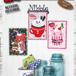 Könnte beinhalten: Präsentation von Mini-Quilts zum Thema Winter, jeweils 15 cm x 23 cm, mit einem Schneemann in einer Tasse, einem Lebkuchenmann und einem Cupcake. Das Bild enthält blaue Einmachgläser, einen Metallbecher und den Text "Winter Nibblers". Knöpfe sind enthalten.