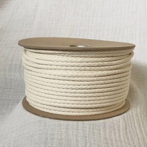 May include: A spool of off-white cotton rope. The rope is tightly wound around a cardboard core. The rope is a natural color and appears to be soft and flexible. The spool is sitting on a white surface.