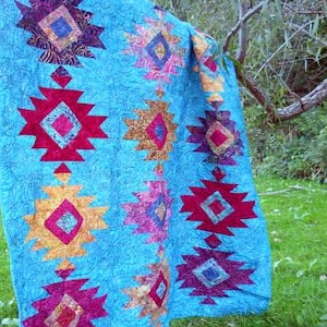 May include: A turquoise quilt with a repeating pattern of geometric shapes in shades of red, gold, purple, and pink. The quilt is hanging outdoors, with a tree branch and green grass in the background.