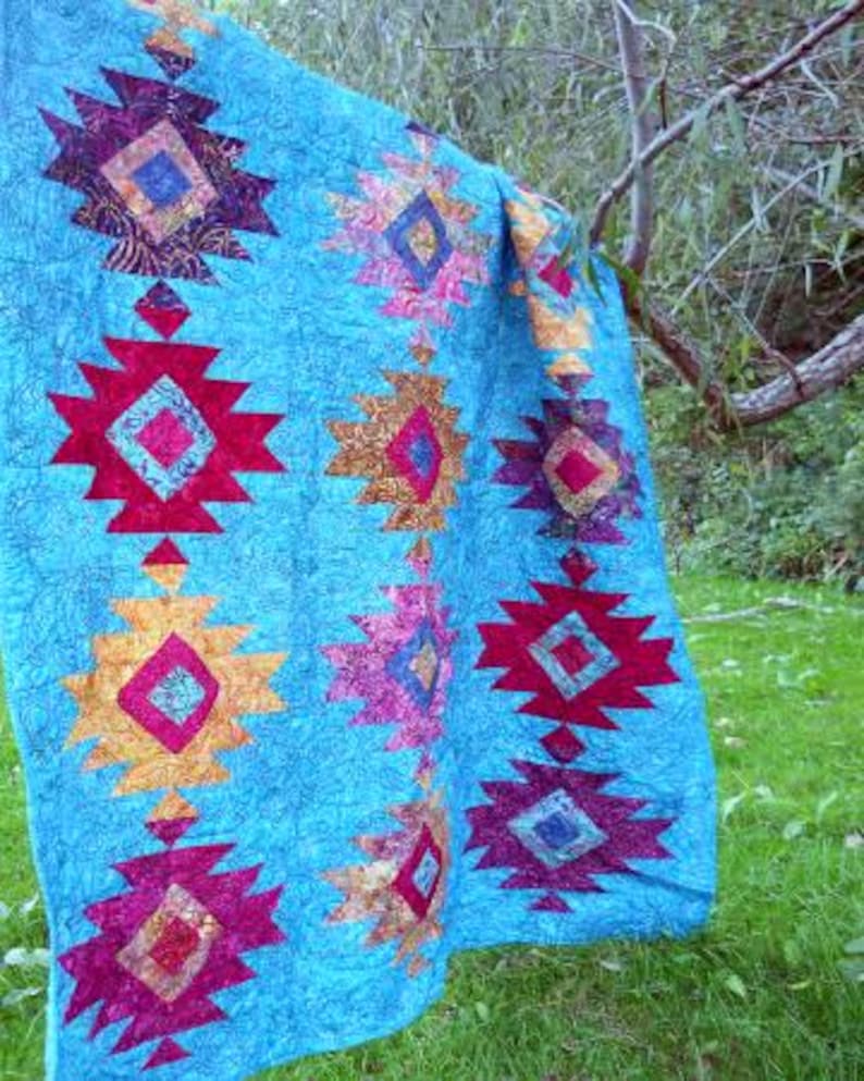May include: A turquoise quilt with a repeating pattern of geometric shapes in shades of red, gold, purple, and pink. The quilt is hanging outdoors, with a tree branch and green grass in the background.