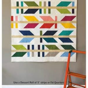 May include: A colorful quilt with arrow designs in various colors, including blue, green, yellow, and red, hangs on a gray wall. The quilt is labeled "The Arrow Maker" and is accompanied by an orange step ladder. The text below reads "Use a Dessert Roll of 5" strips or Fat Quarters."