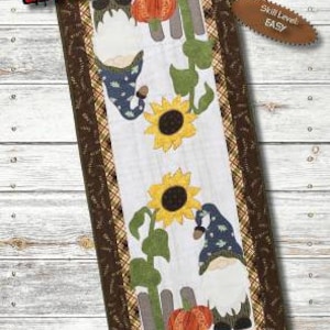May include: A table runner pattern featuring a gnome with a sunflower and pumpkins. The pattern is titled "Home with a Fall Gnome" and is 16 inches by 38 inches. The skill level is easy.