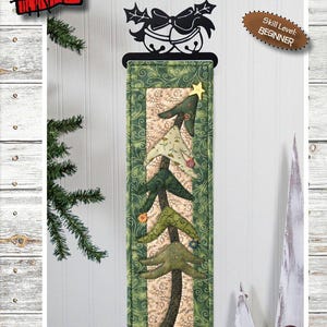 Könnte beinhalten: Ein Wandbehang mit einem Weihnachtsbaum-Design, ca. 15 cm x 51 cm, für Anfänger. Das Design zeigt applizierte Bäume in Grün und Beige, mit einem schwarzen Glocken- und Stechpalmenmotiv oben. Der Text "HOLIDAY TREE" ist zu sehen.