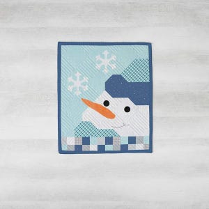 May include: A quilted wall hanging featuring a smiling snowman. The snowman has a carrot nose, black eyes, and a blue hat. The background is light blue with white snowflakes. The quilt is framed with a dark blue border.