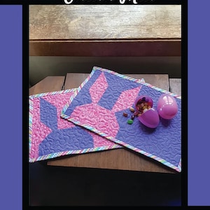 May include: Two purple and pink quilted placemats with a bunny ear design. The placemats are on a wooden table with a pink and white candy dish and two pink plastic eggs.