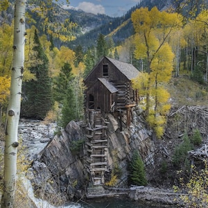 Puede incluir: Una antigua estructura de madera encaramada en un acantilado rocoso, rodeada por un río y árboles. El edificio tiene un aspecto rústico con una escalera que conduce al agua. El paisaje presenta un follaje amarillo vibrante y montañas.