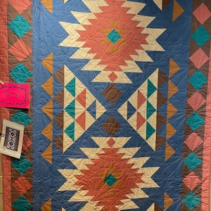 May include: A quilt with a geometric pattern in shades of brown, teal, pink, and white. The quilt is made up of triangles and squares. The quilt is on display with a sign that says "This is a Kit".